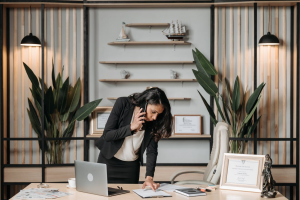A woman standing over a desk reading a document while talking on the phone