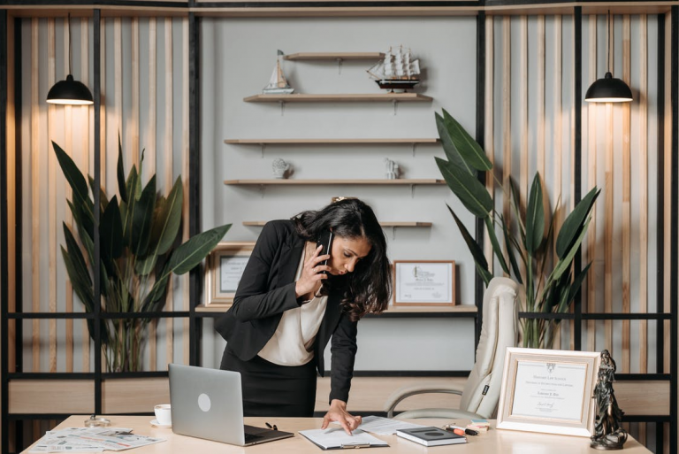 A woman standing over a desk reading a document while talking on the phone