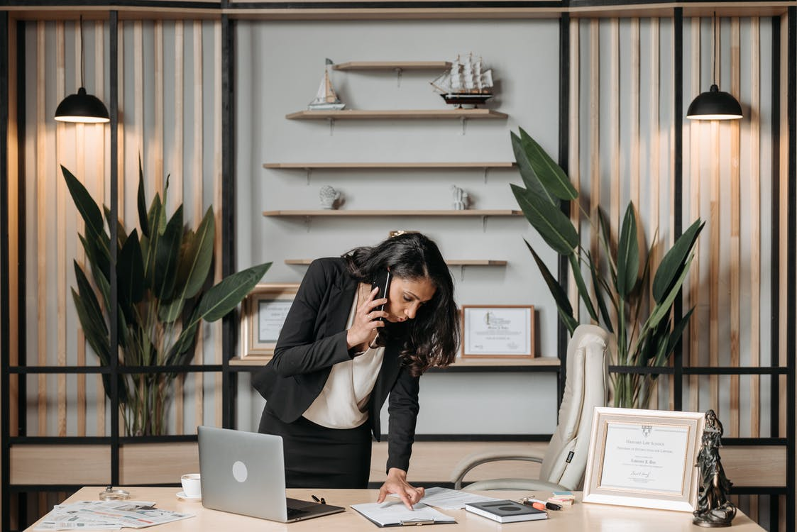 A woman standing over a desk reading a document while talking on the phone